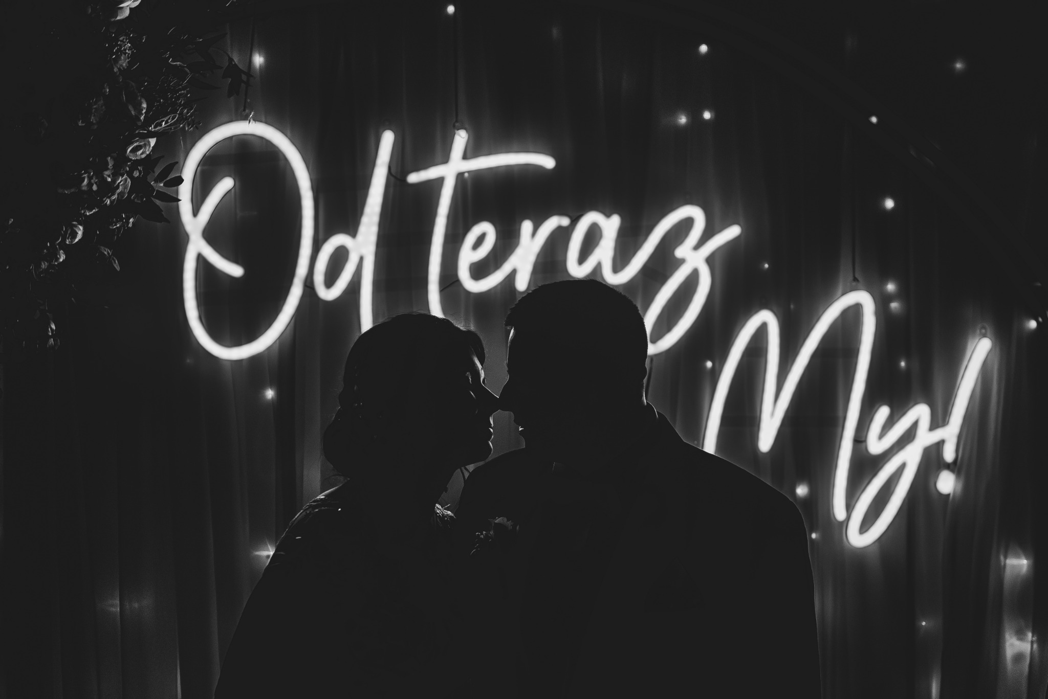 Sylwetki pary młodej w zbliżeniu, na tle neonowego napisu 'Od Teraz My!', dekoracja z kwiatów w tle.