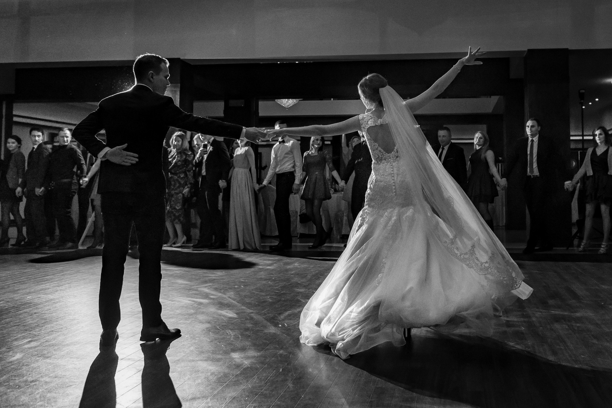Czarno-białe zdjęcie pary młodej podczas pierwszego tańca na parkiecie, z gośćmi w tle, ona w sukni z odkrytymi plecami i długim welonem, on w ciemnym garniturze.