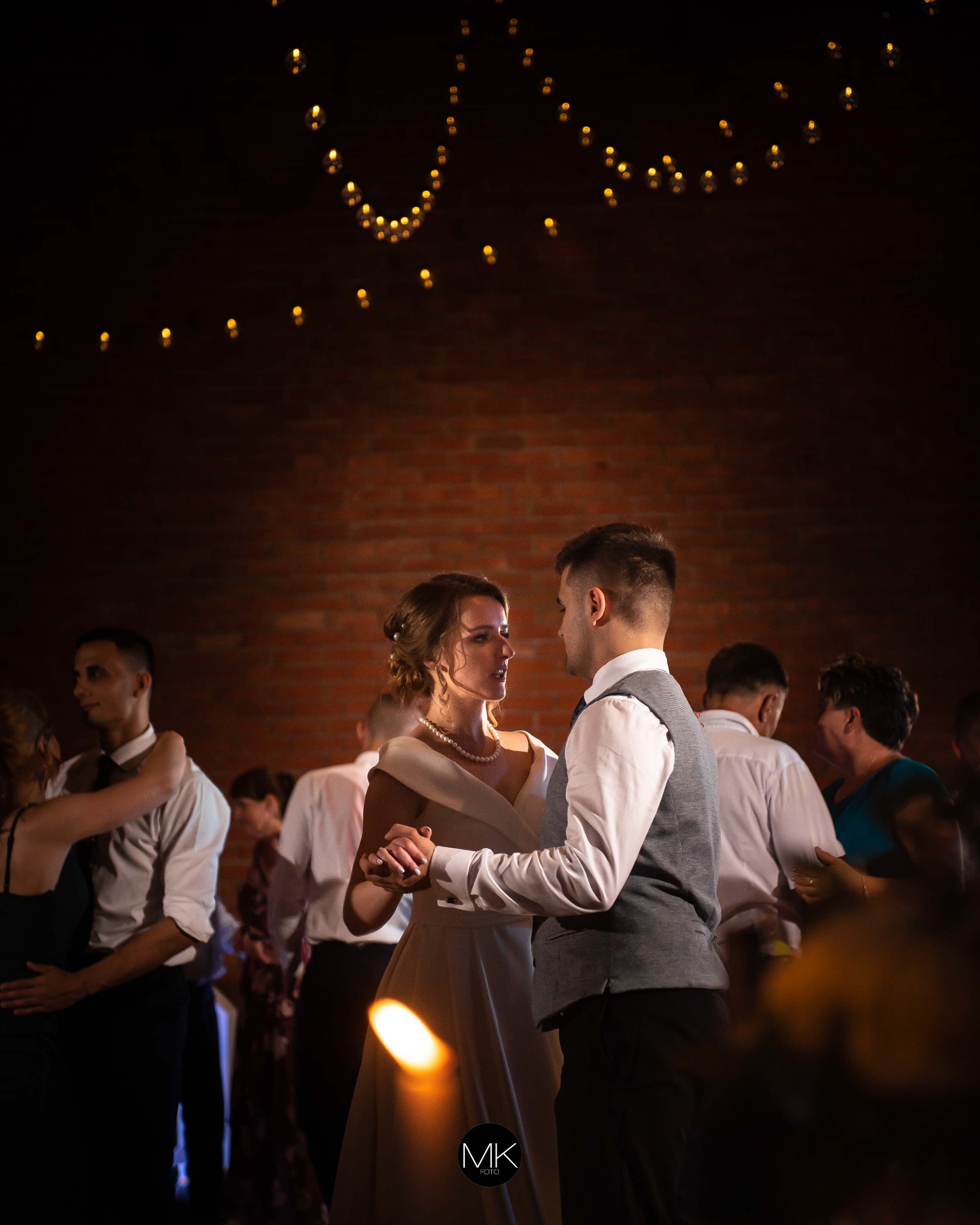 Para młoda tańczy pierwszy taniec na tle ceglanej ściany i girlandy światełek, goście weselni w tle.