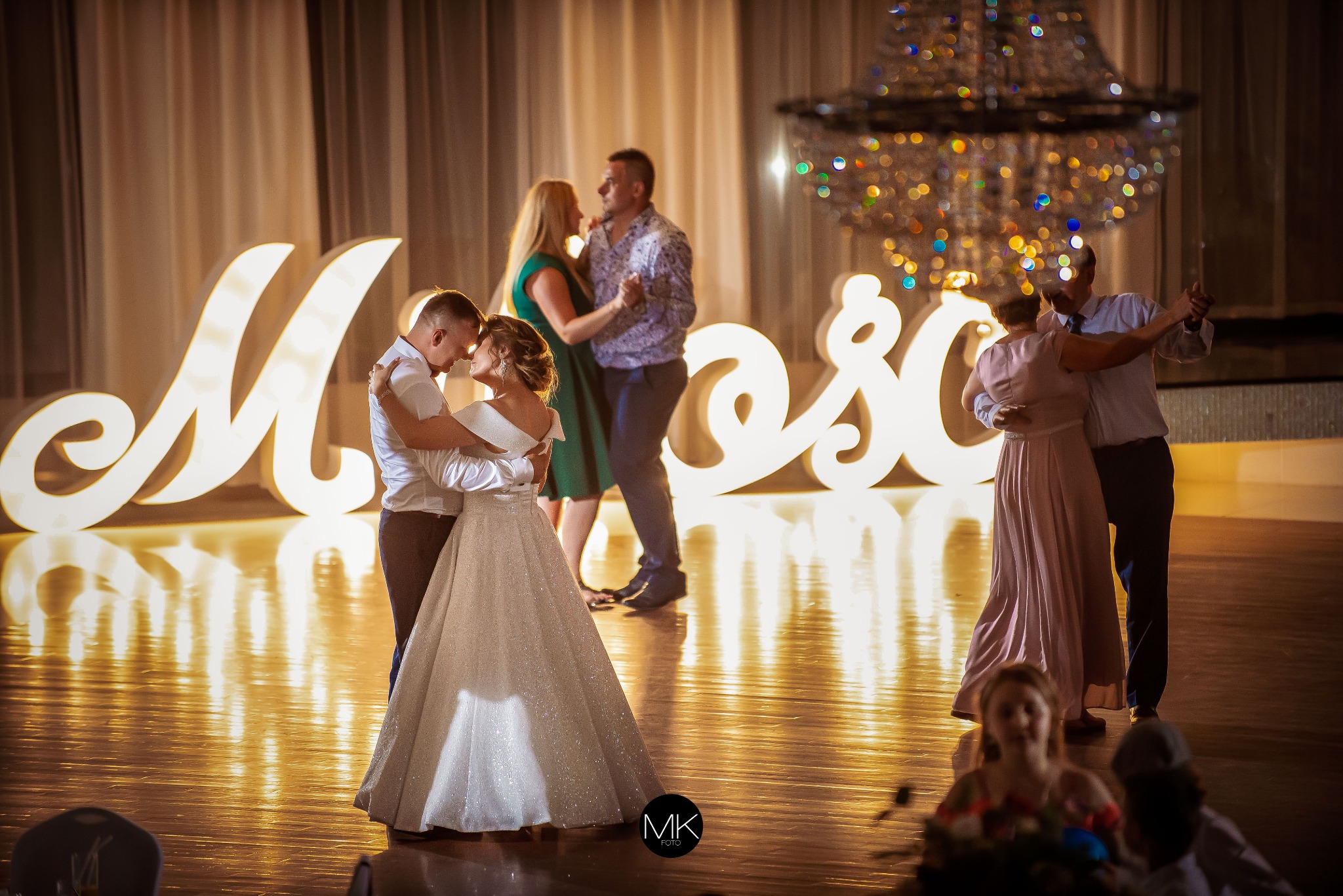 Para młoda tańczy na parkiecie, w tle podświetlane litery, inni goście również tańczą, widoczny żyrandol