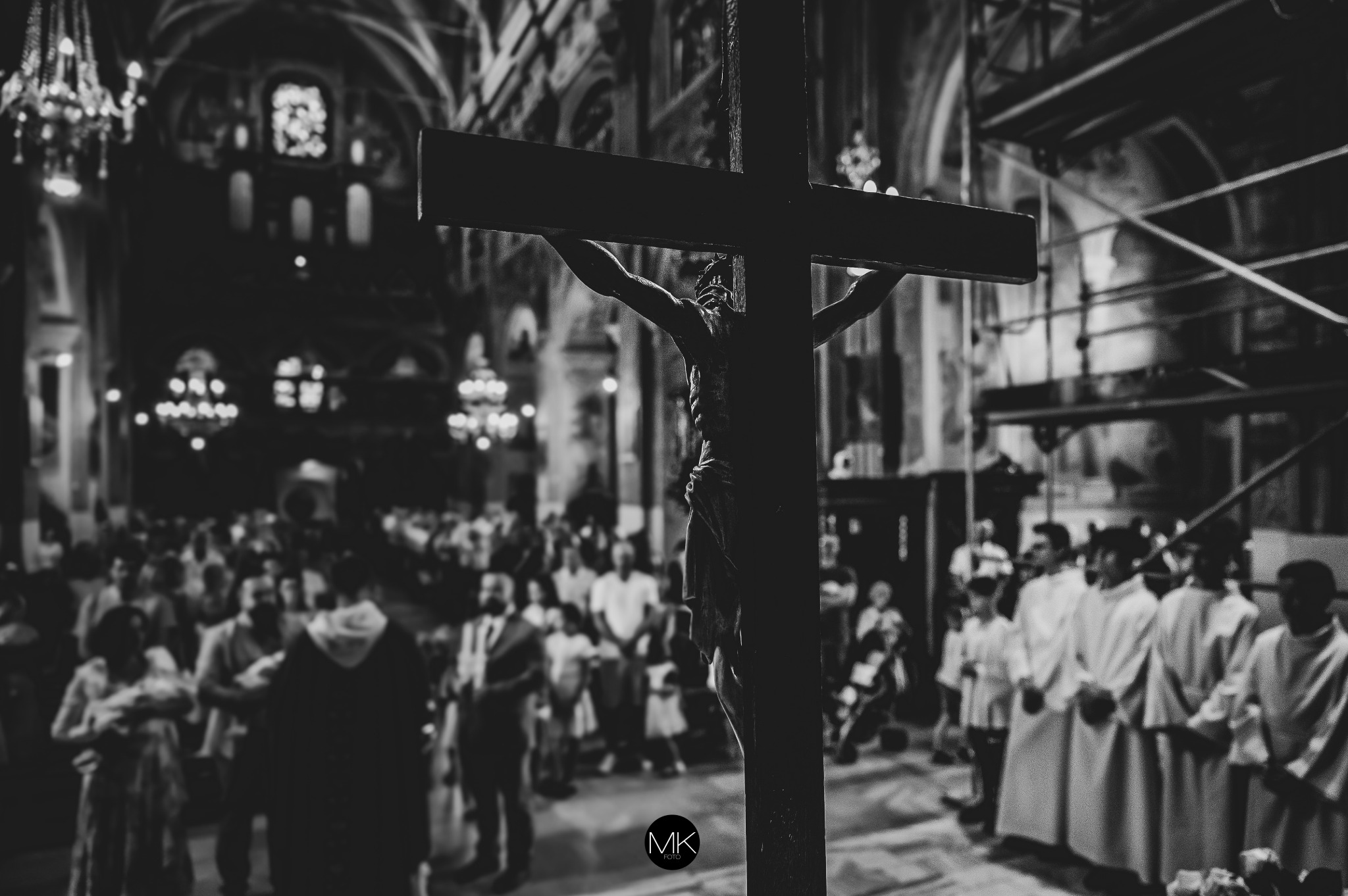 Czarno-białe zdjęcie wnętrza kościoła z krucyfiksem na pierwszym planie i zgromadzonymi ludźmi w tle, oświetlonymi przez żyrandole.