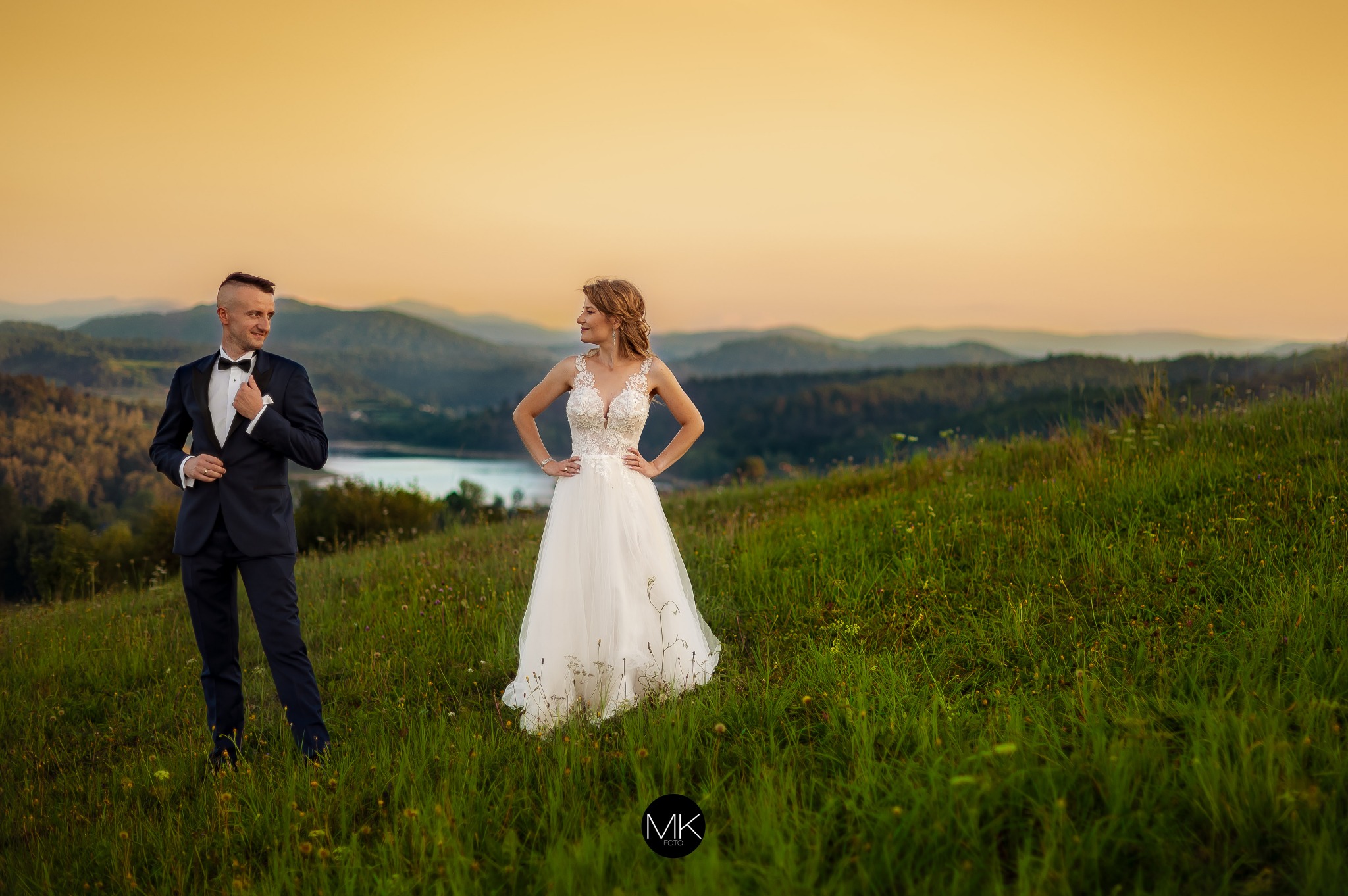 Para młoda w eleganckich strojach pozuje na tle malowniczego krajobrazu z górami i jeziorem podczas zachodu słońca; panna młoda w białej sukni z koronkową górą, pan młody w granatowym garniturze...