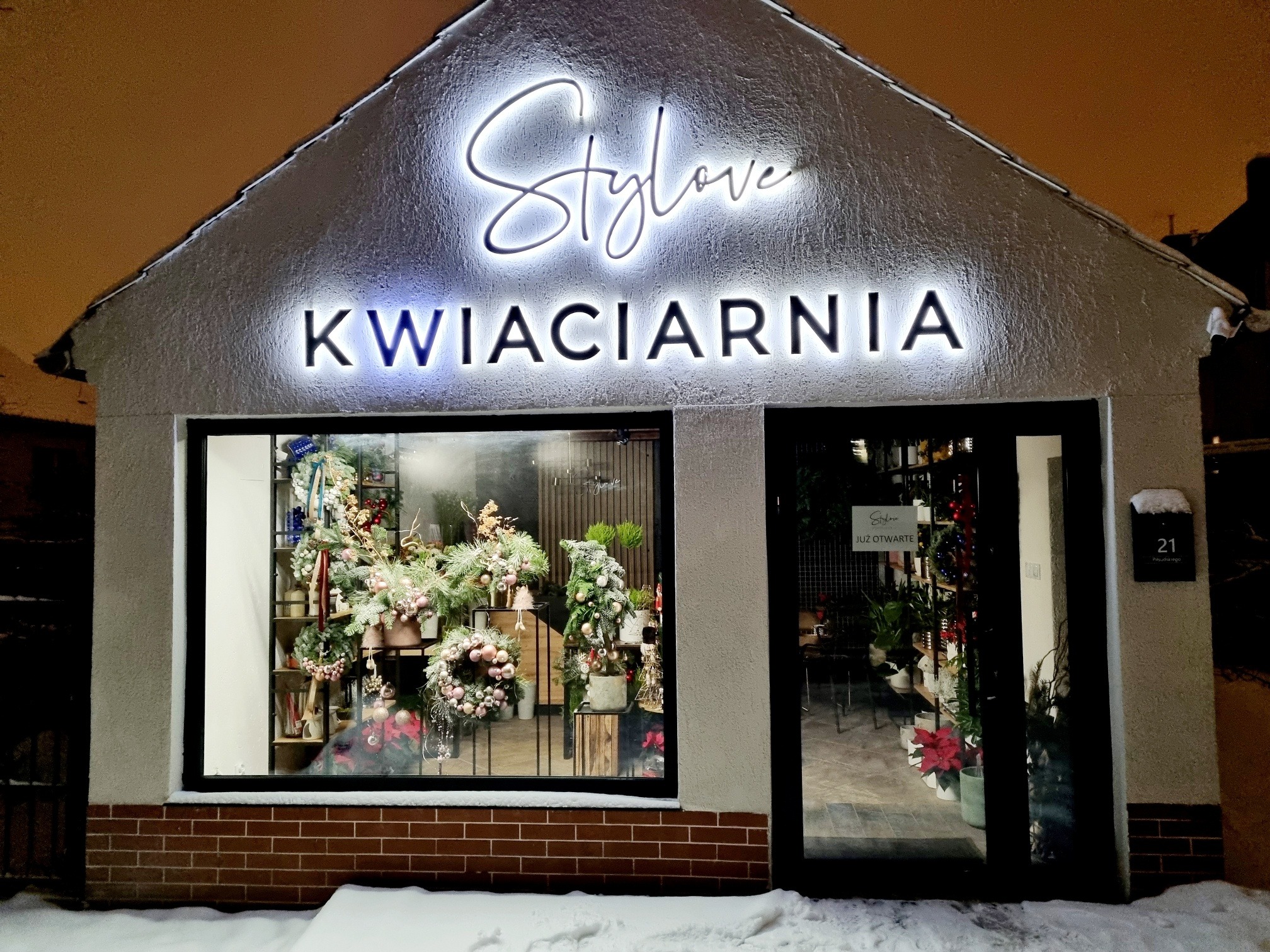 Fasada kwiaciarni 'Stylove' nocą, z podświetlanym neonowym szyldem i świąteczną ekspozycją w oknie, widoczny numer budynku 21 i śnieg na chodniku.