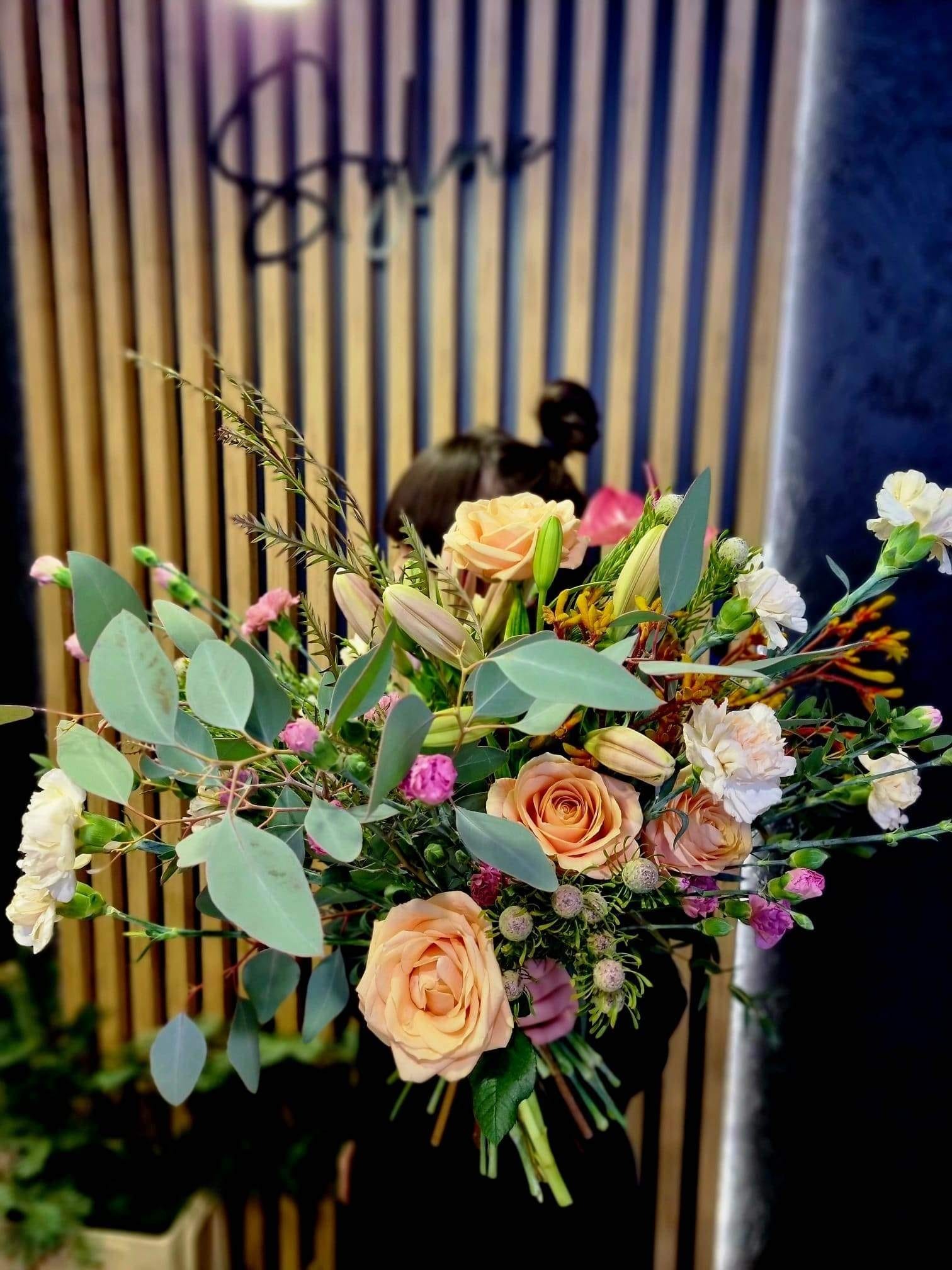 Delikatny bukiet z brzoskwiniowych róż, kremowych goździków i zielonego eukaliptusa na tle drewnianej ściany z pionowymi panelami.