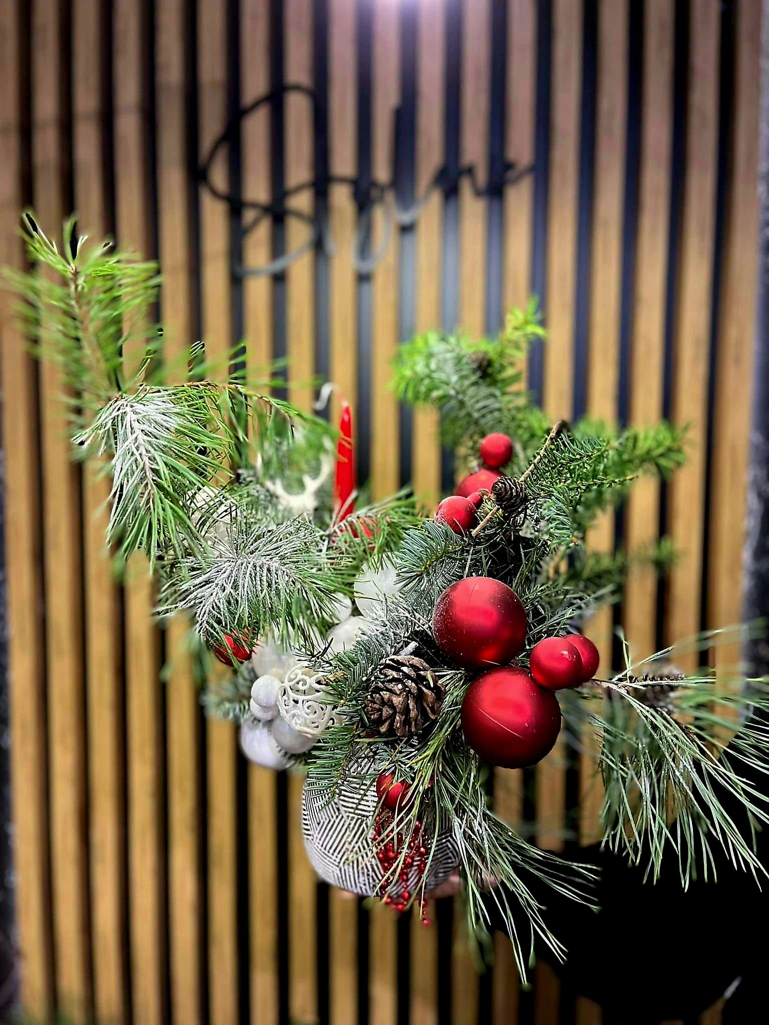 Dekoracja świąteczna z gałązek świerkowych, czerwonych bombek, szyszek i białych ozdób, umieszczona na tle drewnianej ściany z pionowymi listwami.