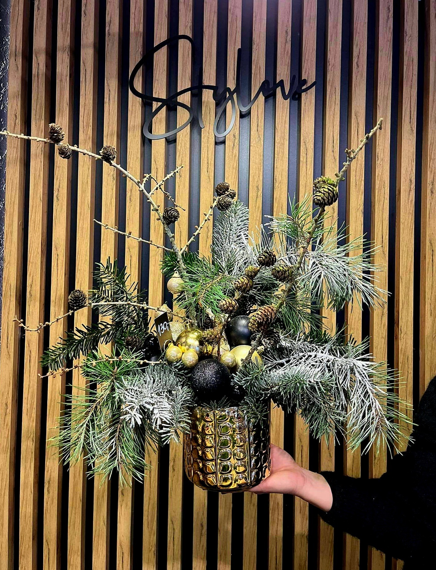 Dekoracja świąteczna z gałązek świerkowych, szyszek i bombek w złotej doniczce na tle drewnianej ściany.