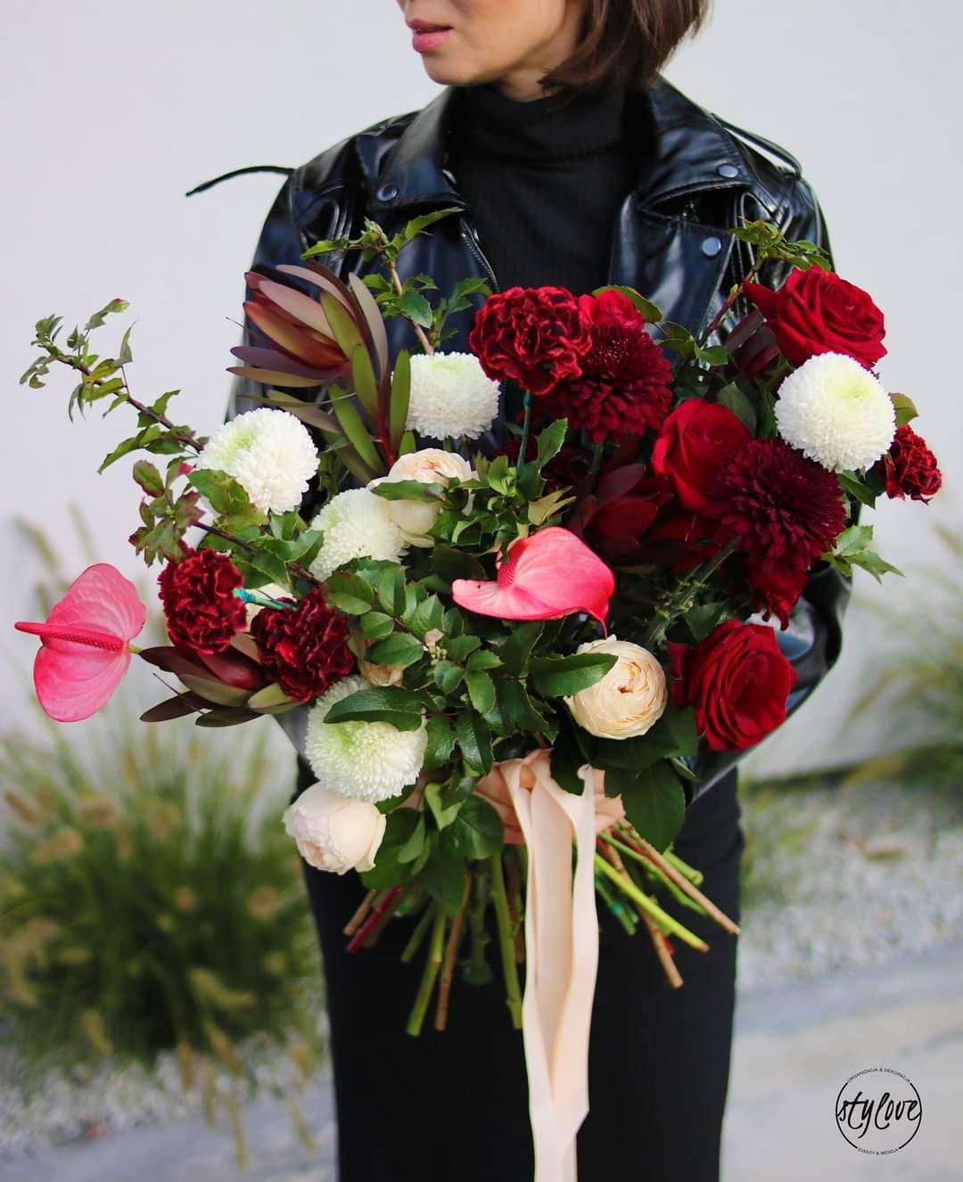 Kobieta w czarnej skórzanej kurtce trzyma w dłoniach duży bukiet czerwono-białych kwiatów z różowym anturium i kremową wstążką.