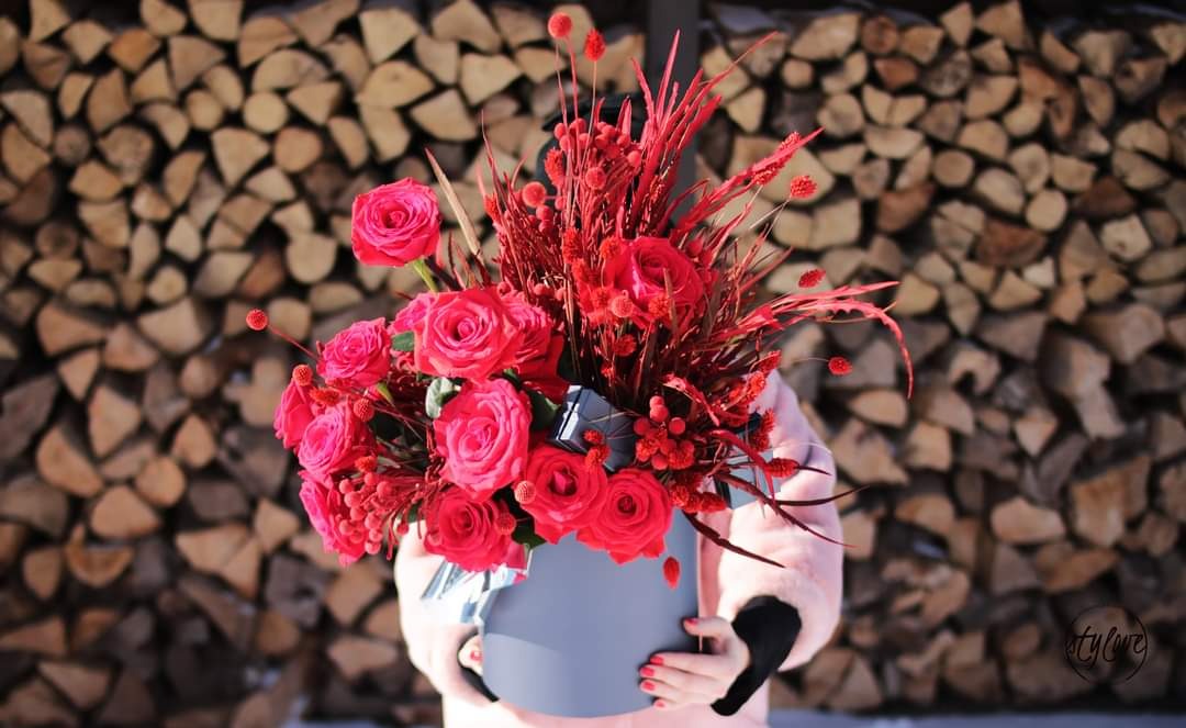 Czerwony bukiet róż w szarym pudełku trzymany przez osobę w różowym futerku na tle stosu drewna.