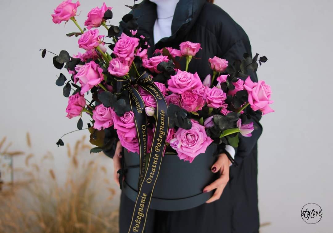 Osoba trzyma w rękach okrągłe pudełko z bukietem różowych róż, czarnych liści eukaliptusa i wstążką z napisem 'Ostatnie Pożegnanie'.