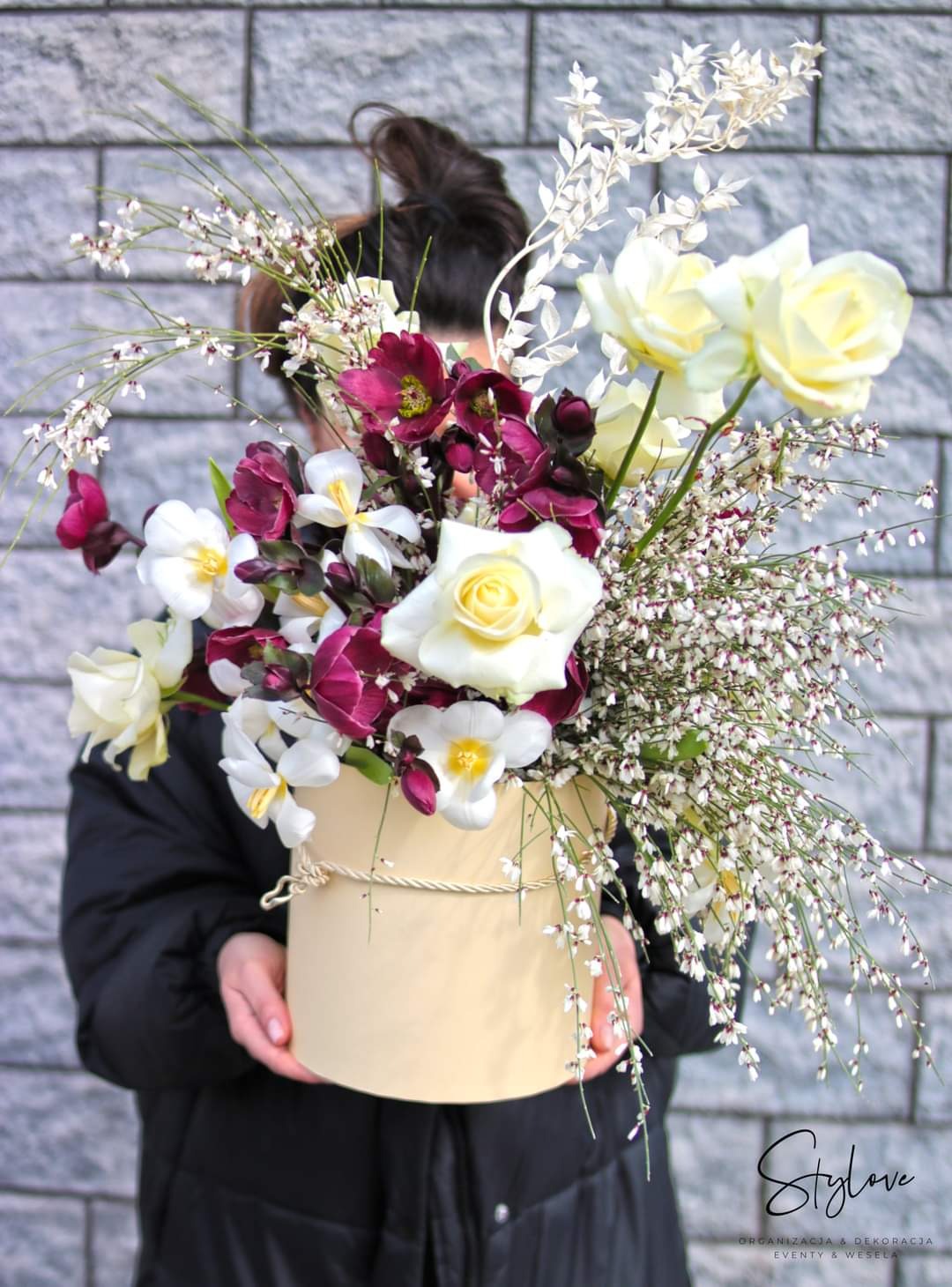 Osoba trzyma w rękach kompozycję kwiatową w okrągłym pudełku, z białymi i bordowymi kwiatami na tle szarej ściany z cegieł.
