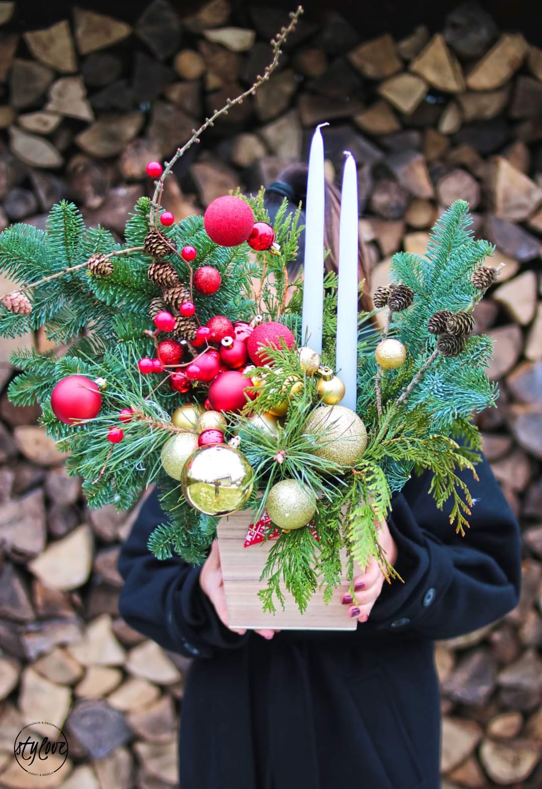 Ozdobny stroik świąteczny z gałązkami świerku, czerwonymi i złotymi bombkami, szyszkami i białymi świecami, trzymany przez osobę na tle ułożonego drewna, wykonany w Olsztynie.
