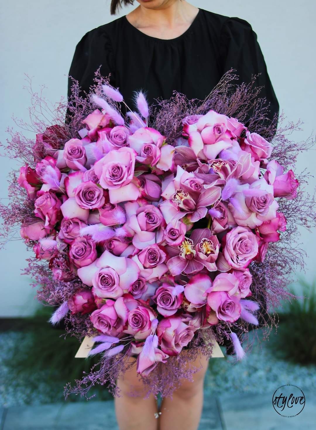 Duży bukiet w kształcie serca z różowych róż i orchidei, ozdobiony drobnymi piórkami i gałązkami, trzymany przez osobę w czarnej sukience, na jasnym tle.