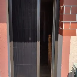 Zewnętrzna moskitiera plisowana w kolorze antracytowym, zamontowana w drzwiach wejściowych, widok z zewnątrz na fragment imitacji trawy i klimatyzator.