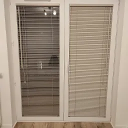 Białe, dwuskrzydłowe drzwi balkonowe z zamontowanymi poziomymi żaluzjami w kolorze beżowym, widoczne z bliska na tle drewnopodobnej podłogi.