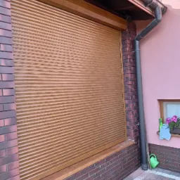Zasłonięte brązowe okno roletą zewnętrzną w Kraśniku, widoczny fragment elewacji z cegły i rynna spustowa.