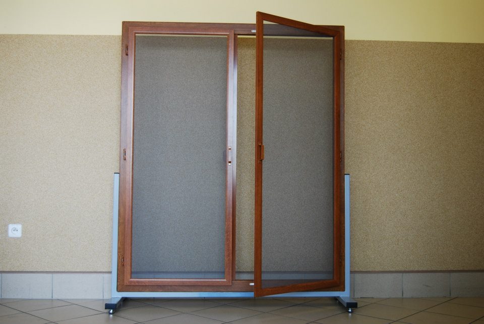 Brązowa rama okienna z szarym wypełnieniem, jedna część otwarta, prezentowana na metalowym stojaku wewnątrz pomieszczenia z beżową ścianą i płytkami podłogowymi.