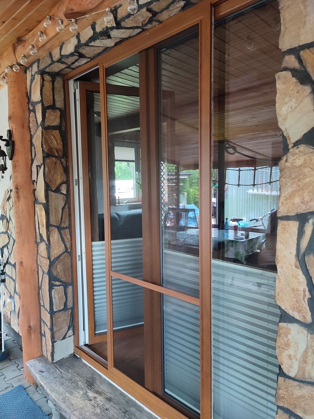Zewnętrzny widok przeszklonego, brązowego okna przesuwnego z częściowo zasłoniętymi plisowanymi roletami, kamienna elewacja budynku i drewniany sufit werandy.