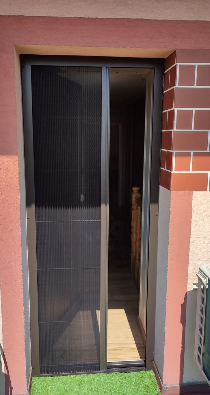 Zewnętrzna moskitiera plisowana w kolorze antracytowym, zamontowana w drzwiach wejściowych, widok z zewnątrz na fragment imitacji trawy i klimatyzator.