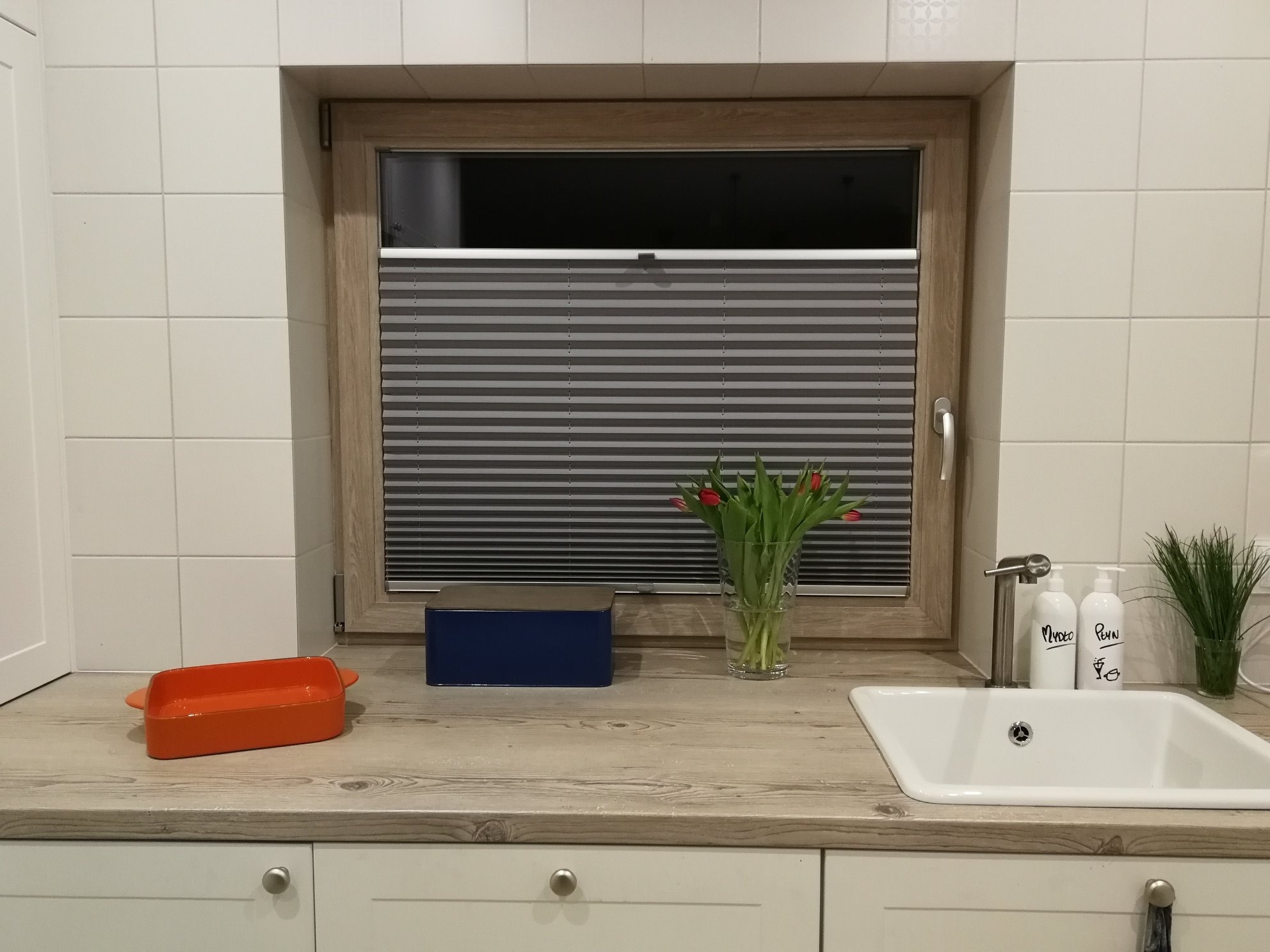 Kuchnia z oknem, w którym zamontowane są szare, plisowane przesłony okienne. Na blacie stoją tulipany w wazonie, niebieskie pudełko i pomarańczowa forma do pieczenia. Nad zlewem widoczne dozowniki...