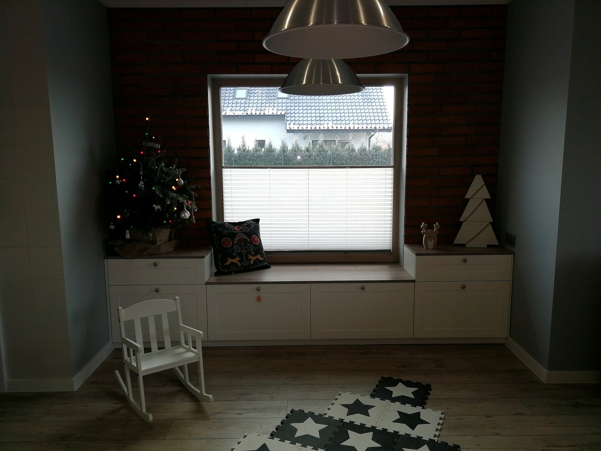 Pokój z ceglaną ścianą, oknem z białymi plisami, zabudową meblową pod oknem, choinką i dekoracjami świątecznymi.