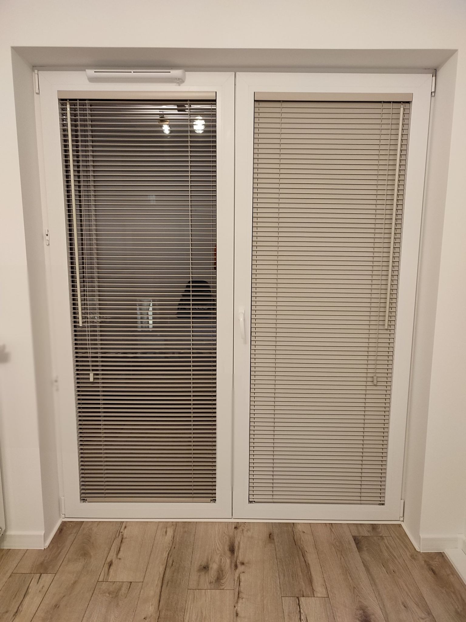 Białe, dwuskrzydłowe drzwi balkonowe z zamontowanymi poziomymi żaluzjami w kolorze beżowym, widoczne z bliska na tle drewnopodobnej podłogi.