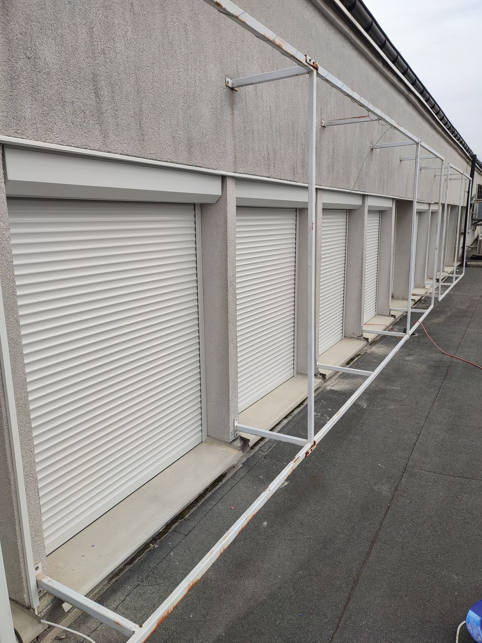 Szereg białych rolet zewnętrznych, zamkniętych, zamontowanych w budynku o szarej, chropowatej elewacji, z widoczną metalową konstrukcją rusztowania. Widok z zewnątrz, z poziomu dachu.