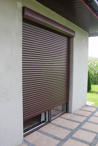 Brązowa roleta zewnętrzna częściowo opuszczona na oknie tarasowym, widoczne fragmenty ogrodu i kostki brukowej na tarasie.