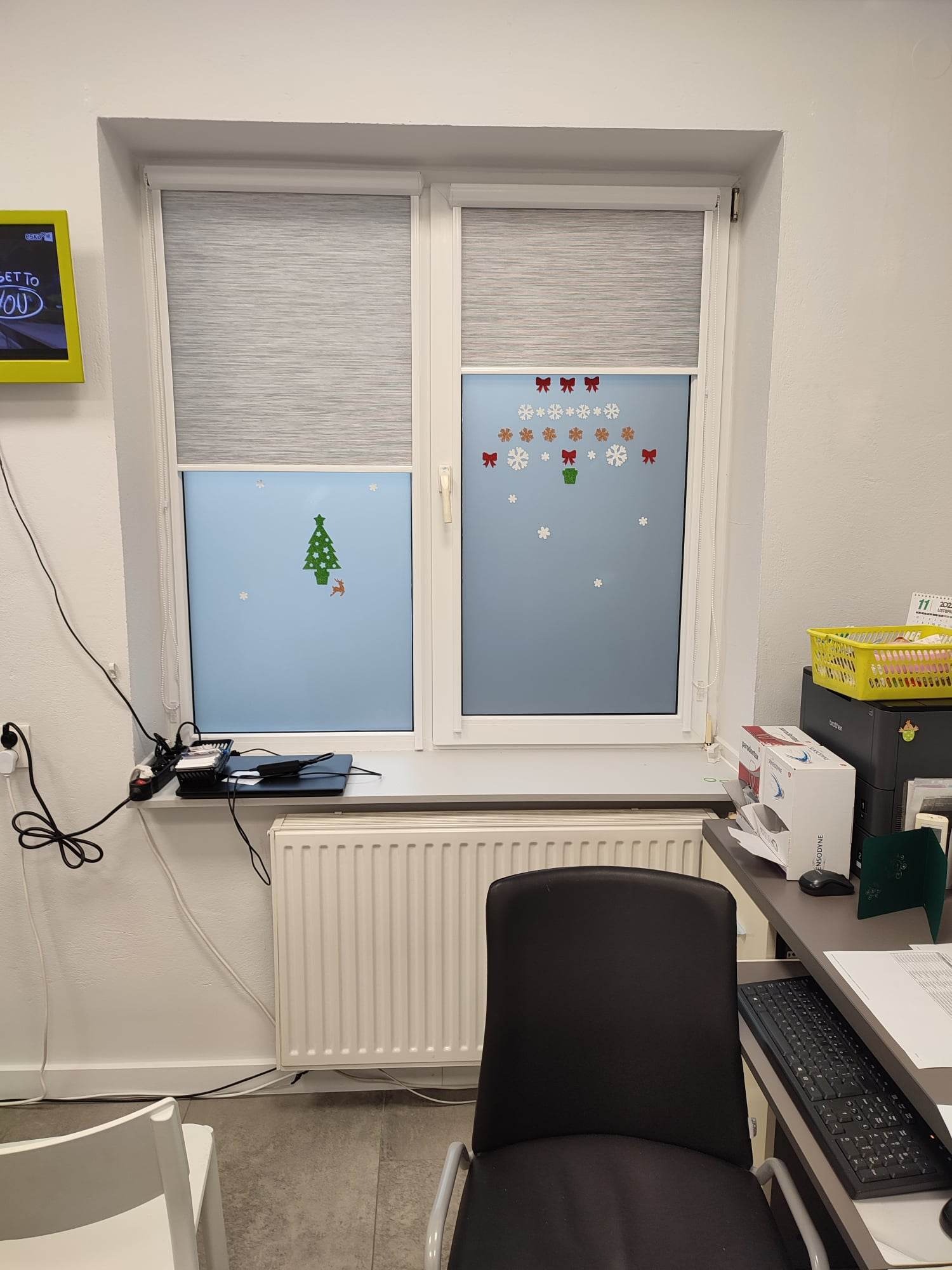 Dwa okna z częściowo zasłoniętymi roletami, ozdobione świątecznymi naklejkami, widok z wnętrza biura z biurkiem i sprzętem biurowym.
