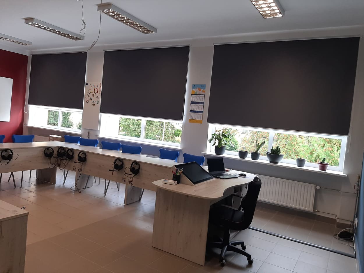 Zasłonięte rolety wewnętrzne w sali lekcyjnej z biurkami, krzesłami i roślinami na parapecie.