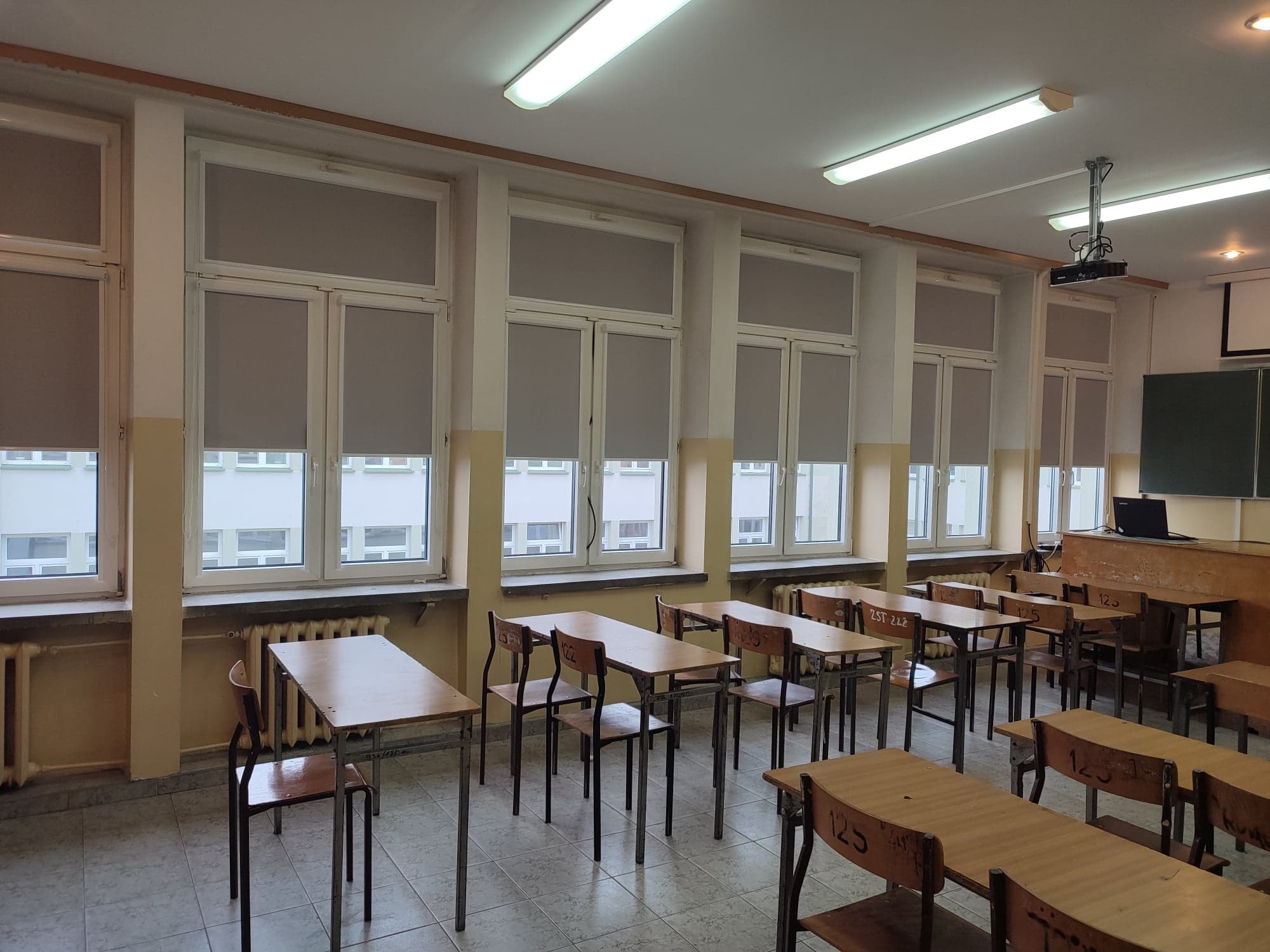Wnętrze klasy szkolnej z rzędami drewnianych ławek i krzeseł, okna z szarymi roletami w pozycji zamkniętej, rzutnik na suficie.
