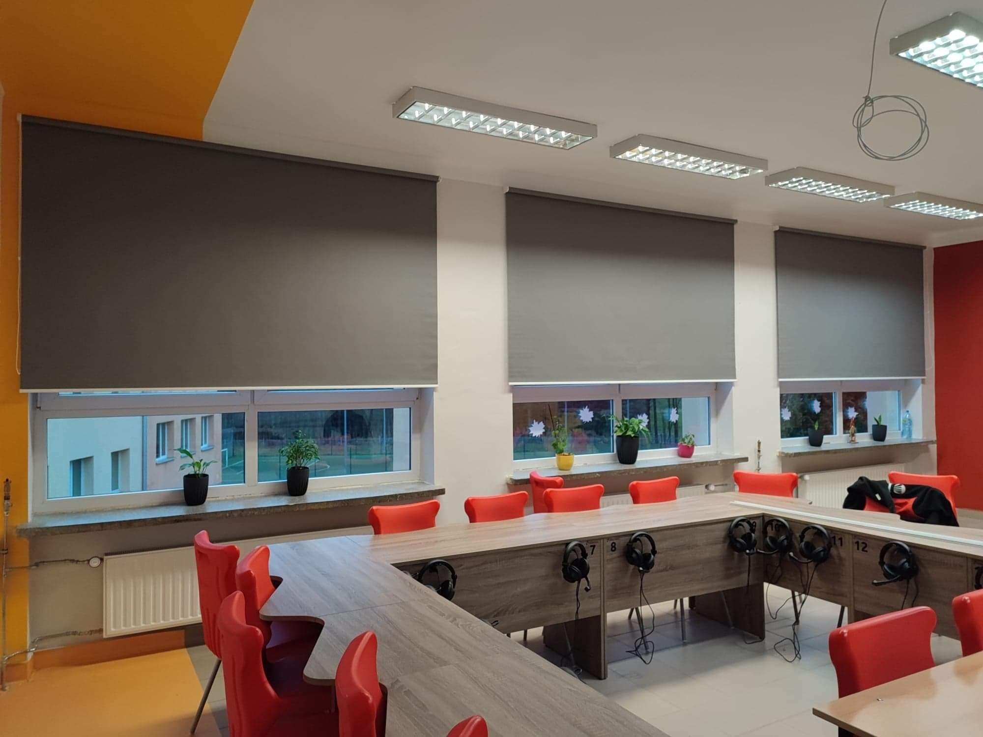 Wnętrze sali lekcyjnej z trzema oknami wyposażonymi w szare rolety zwinięte do połowy, czerwone krzesła i stoły z przymocowanymi słuchawkami.