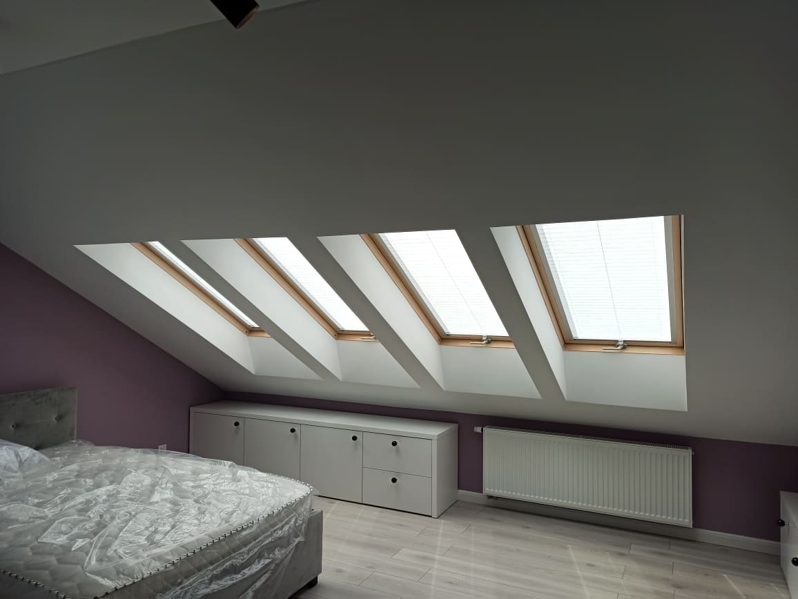 Poddasze z czterema oknami dachowymi wyposażonymi w białe żaluzje poziome, widoczne łóżko z materacem w folii ochronnej i białe meble.