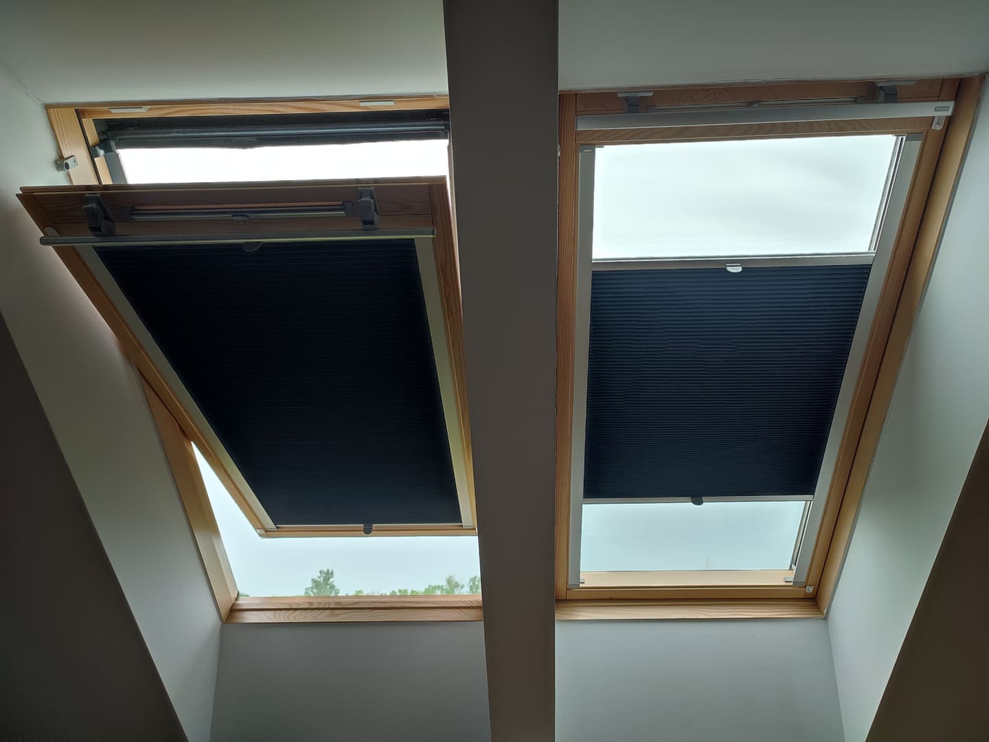 Dwa okna dachowe z drewnianą ramą, jedno z częściowo opuszczoną, plisowaną, zaciemniającą roletą w kolorze czarnym, drugie z całkowicie zasłoniętą roletą, widok z dołu.