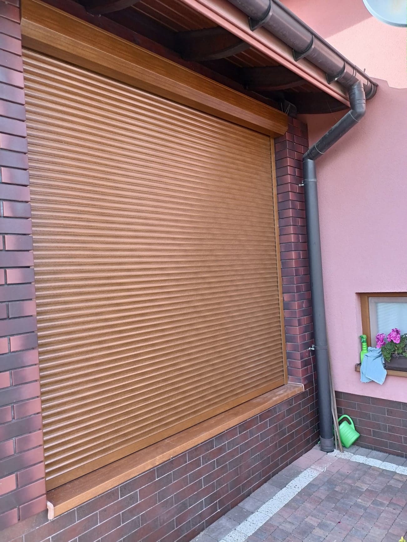 Zasłonięte brązowe okno roletą zewnętrzną w Kraśniku, widoczny fragment elewacji z cegły i rynna spustowa.