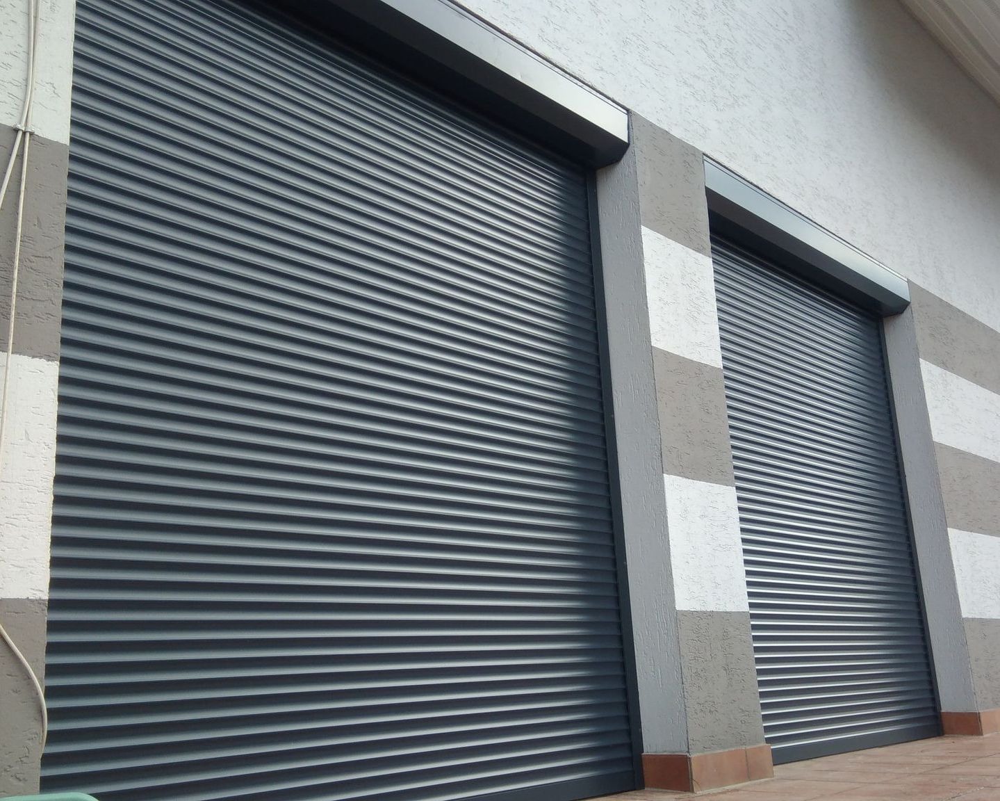 Zamknięte, szare rolety antywłamaniowe zamontowane w elewacji budynku z dekoracyjnymi pasami w odcieniach szarości i bieli.