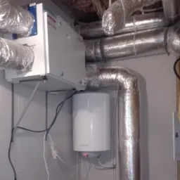 Zamontowany rekuperator z widocznymi przewodami wentylacyjnymi w pomieszczeniu z surowym wykończeniem, widoczny biały zbiornik wyrównawczy.