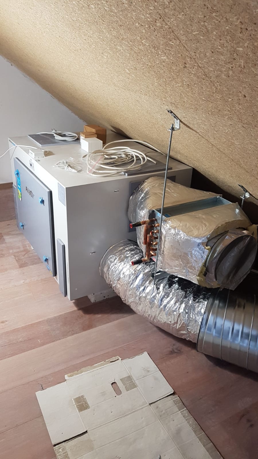 Jednostka rekuperacyjna w szarej obudowie z podłączonymi przewodami wentylacyjnymi, zainstalowana na poddaszu o skośnych ścianach i drewnianej podłodze. Widoczne kable i akcesoria na górnej...