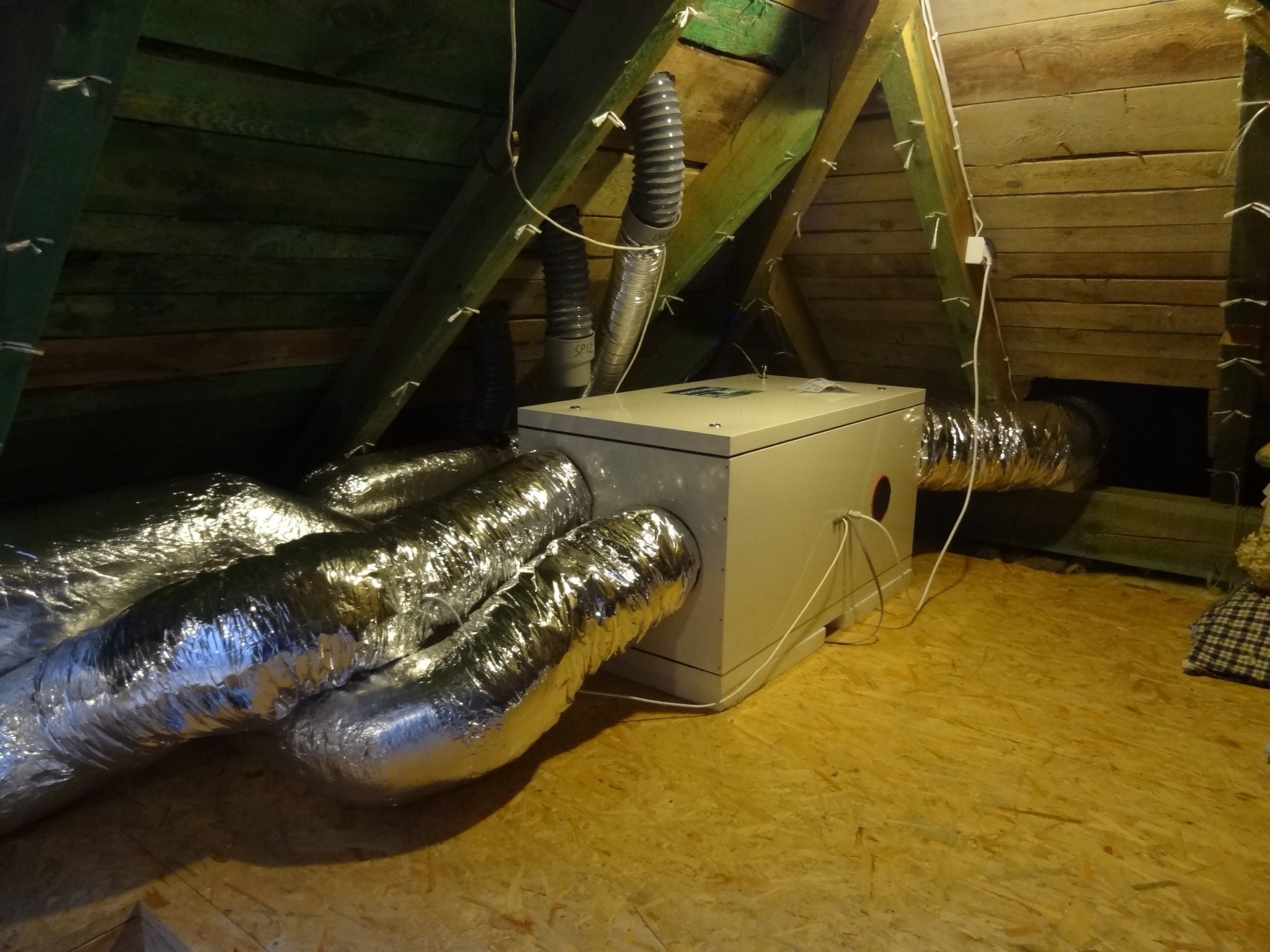 Centrala wentylacyjna z podłączonymi, błyszczącymi przewodami wentylacyjnymi w przestrzeni poddasza z widoczną drewnianą konstrukcją dachu.