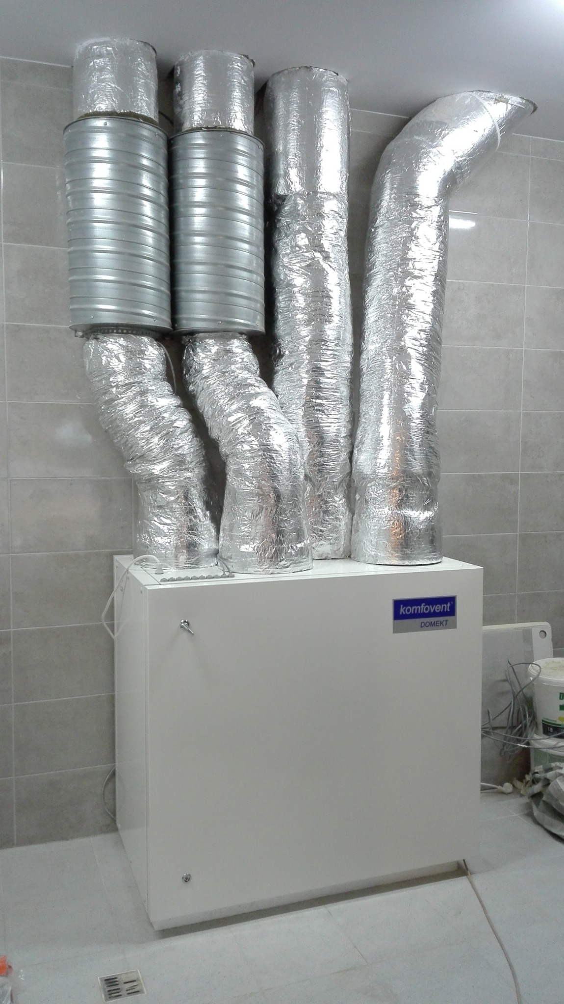Centrala wentylacyjna Komfovent Domekt z podłączonymi przewodami wentylacyjnymi, niektóre owinięte folią aluminiową, zamontowana w pomieszczeniu z szarymi płytkami.