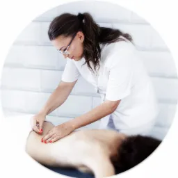 Kobieta w białym uniformie i okularach aplikuje igły na plecy pacjentki leżącej na stole do masażu w jasnym pomieszczeniu z białymi kafelkami w tle.