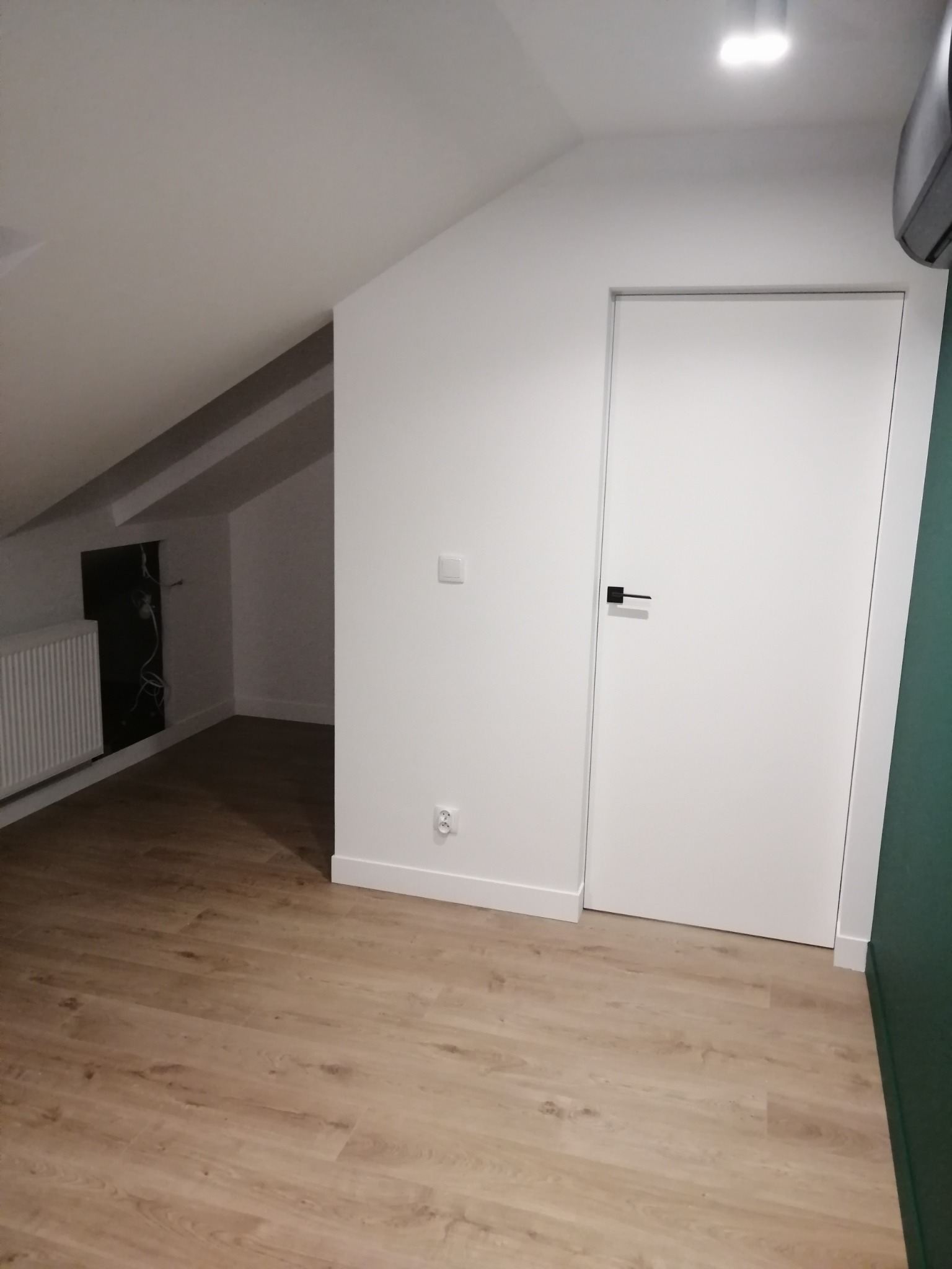 Jasne panele podłogowe ułożone w pokoju na poddaszu, białe ściany z widocznymi gniazdkami i włącznikami, białe drzwi z czarną klamką, grzejnik i instalacja elektryczna wnęce.