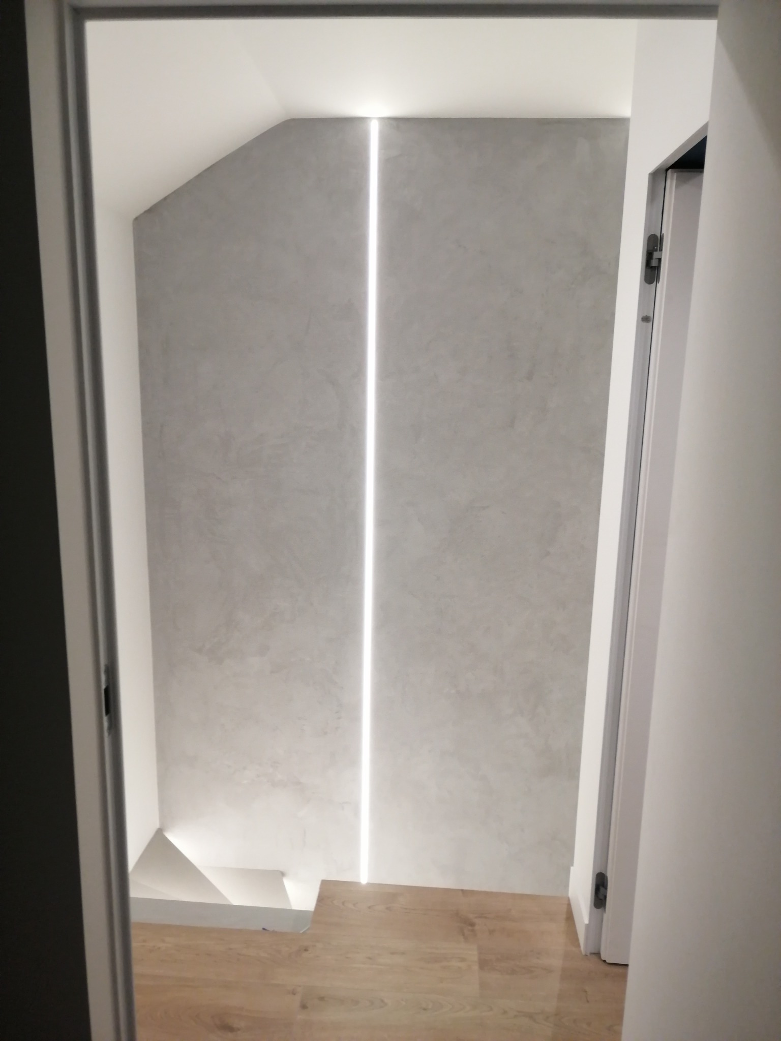 Nowoczesne wykończenie wnętrza z pionowym oświetleniem LED w ścianie o teksturze betonu, widoczne schody i drewniana podłoga