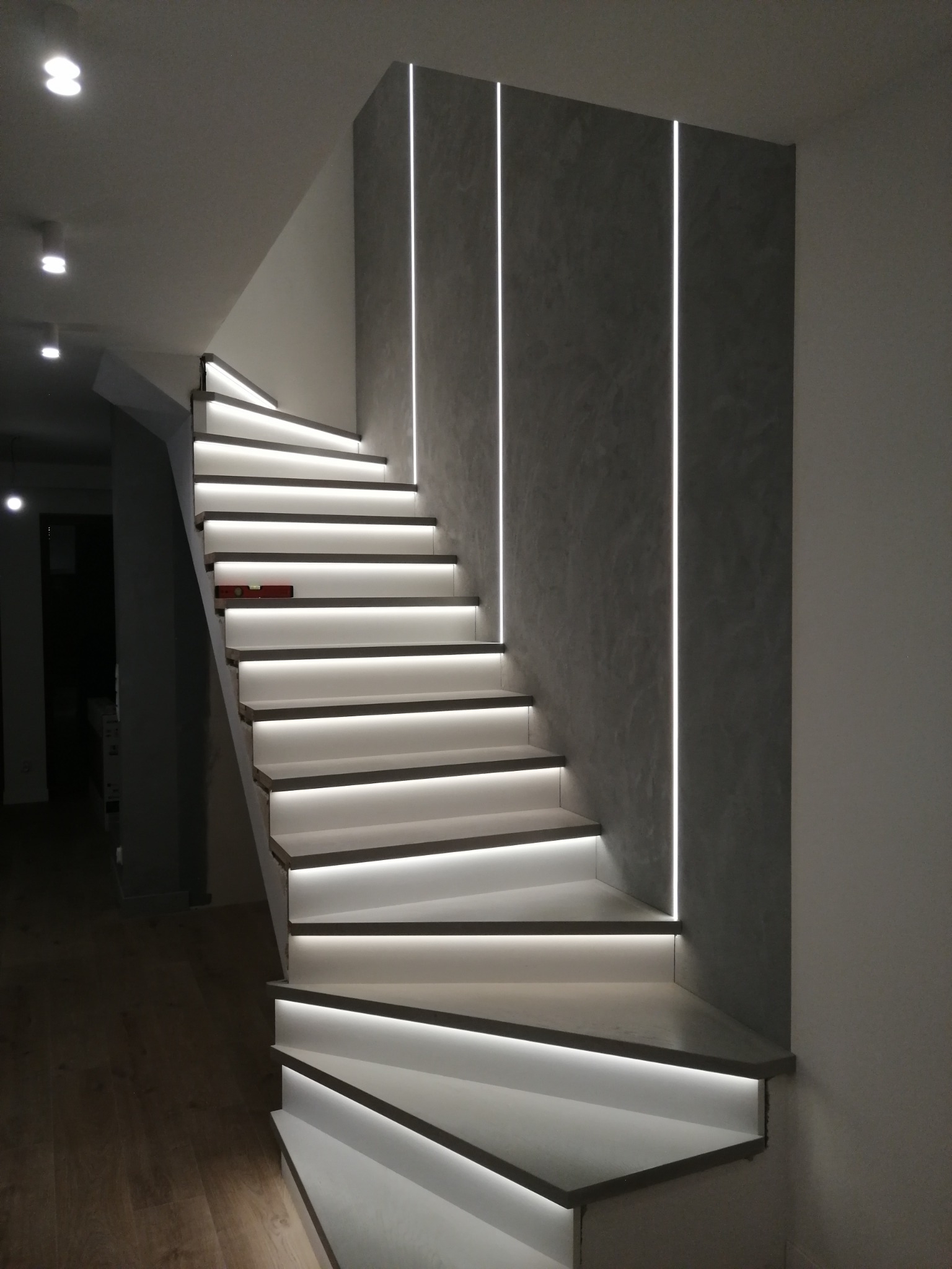 Nowoczesne schody z podświetleniem LED na każdym stopniu, ściana z szarego betonu architektonicznego z pionowymi wpuszczonymi paskami LED, poziomica na czwartym stopniu od góry.