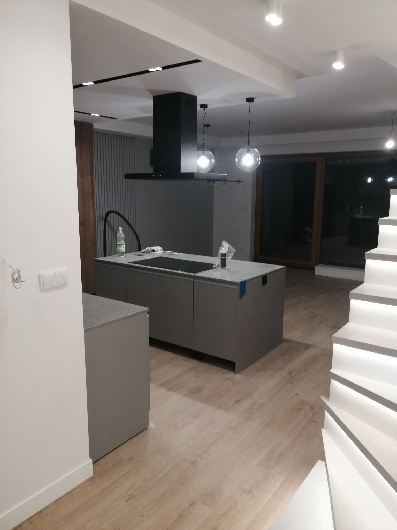 Nowoczesna szara kuchnia z wyspą, czarnym okapem i oświetleniem punktowym, widok na podświetlane schody i drewnianą podłogę.