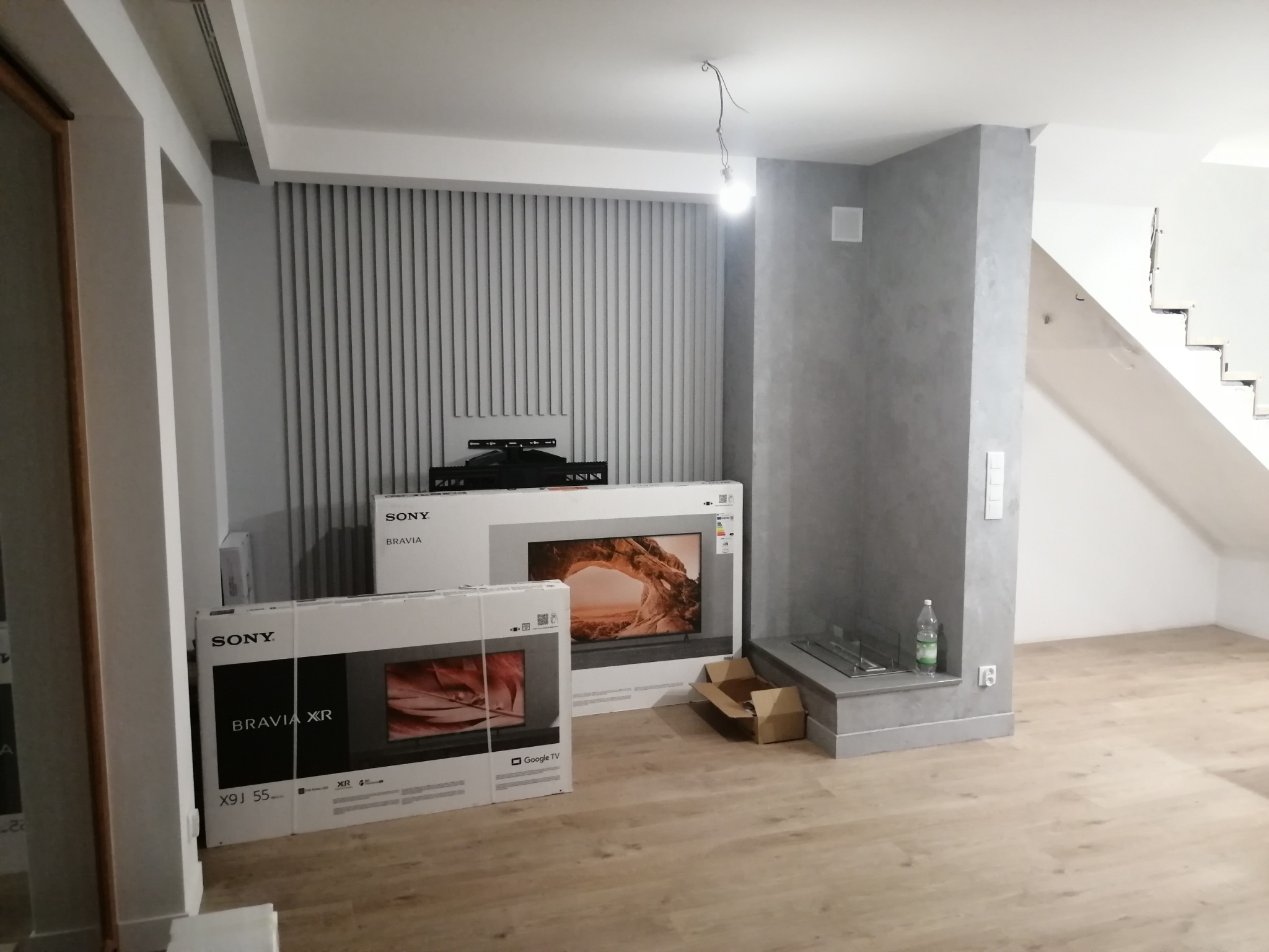 Salon w trakcie urządzania: dwa telewizory Sony Bravia XR w kartonach na podłodze z paneli, ściana z szarych pionowych paneli dekoracyjnych i kominek na biopaliwo w szarej obudowie.