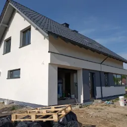 Nowoczesny dom jednorodzinny z białą elewacją i grafitową stolarką okienną, widok z zewnątrz na częściowo wykończony budynek z paletami i workami budowlanymi w tle.