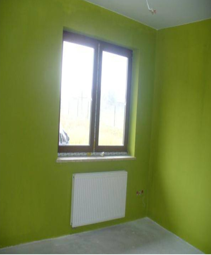 Pokój po malowaniu ścian na zielono, z oknem i zamontowanym grzejnikiem, widoczne niedoskonałości wykończenia przy suficie.