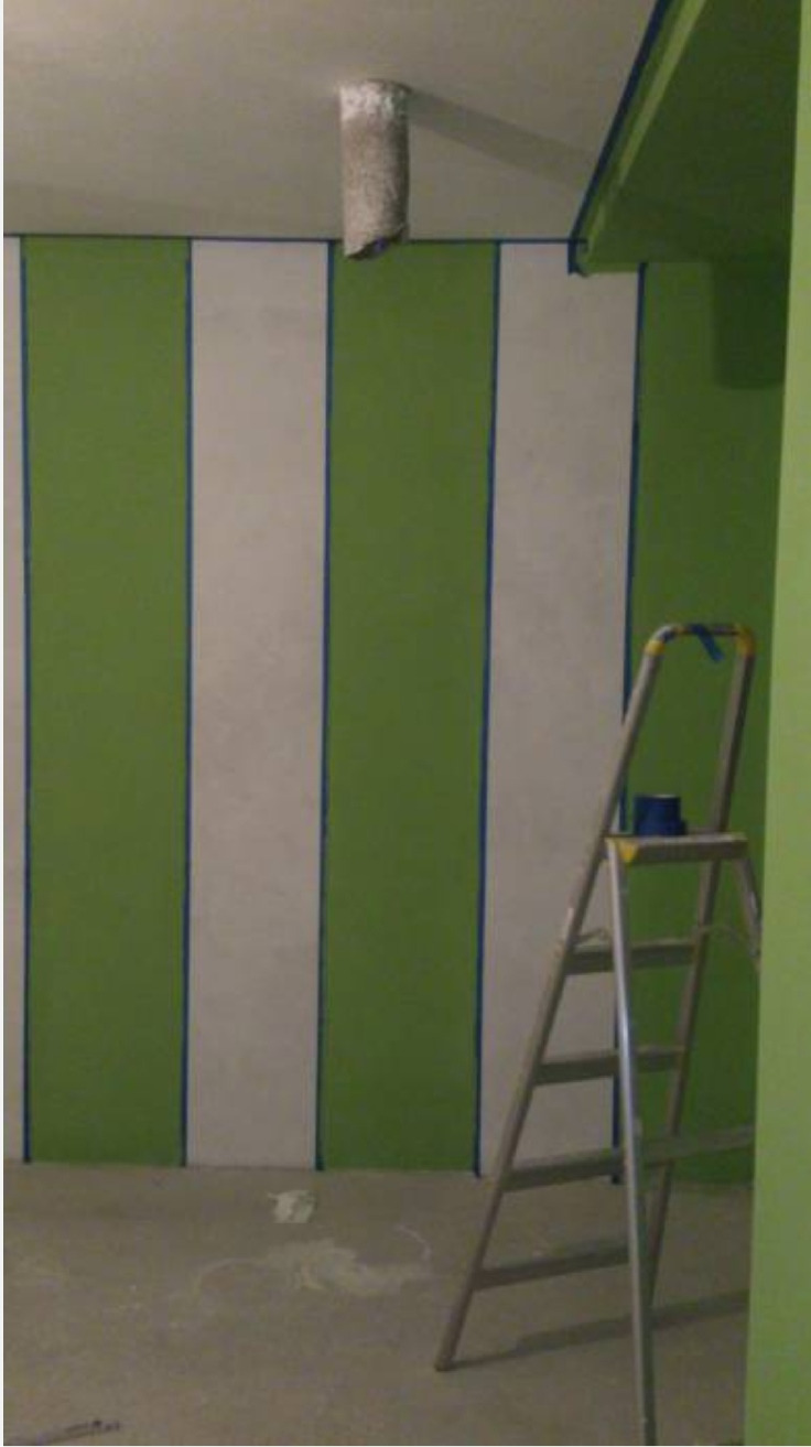 Malowanie ścian w pionowe pasy zieleni i bieli, zabezpieczone niebieską taśmą malarską, drabina aluminiowa i tuba wentylacyjna wystająca z sufitu.