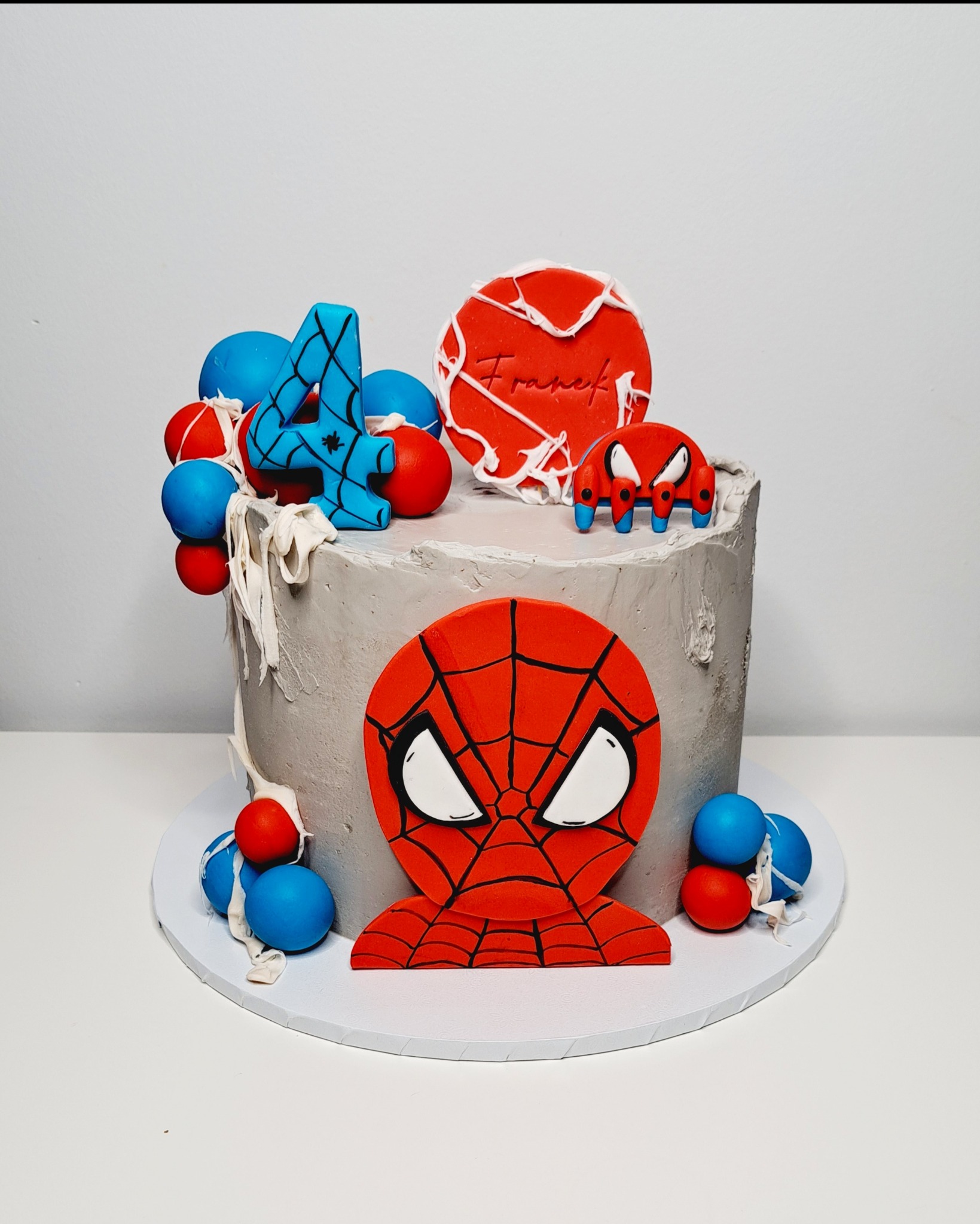 Tort urodzinowy w stylu Spider-Mana z szarym kremem, dekoracjami z masy cukrowej: głowa Spider-Mana, liczba 4, czerwony okrąg z napisem 'Franek' i mały pająk, otoczony niebiesko-czerwonymi kulkami.