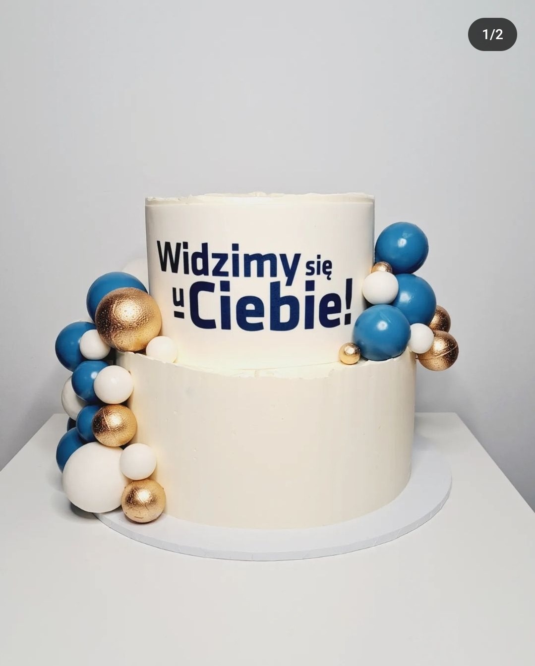 Dwupoziomowy tort z białym kremem, ozdobiony niebieskimi, złotymi i białymi kulkami oraz napisem 'Widzimy się u Ciebie!' w kolorze niebieskim.